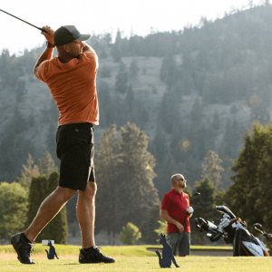 Man dressed in orange shirt and shorts swings a golf club while his friend looks on.